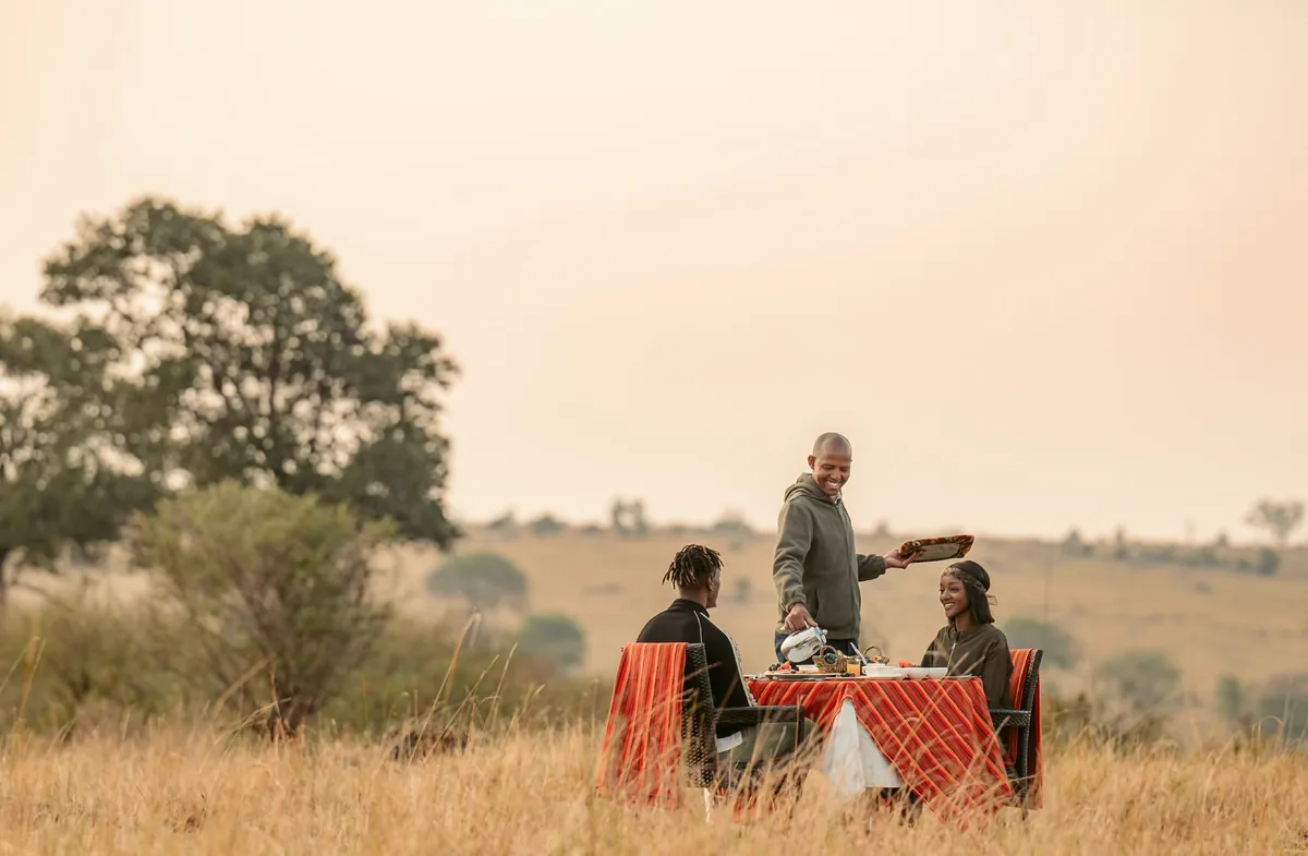 A waiter serving breakfast to two guests at a table set out in the long grass at dawn