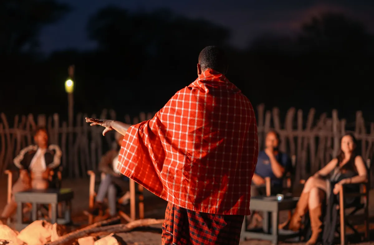 A Maasai storyteller in a red shuka speaking to guests seated around the evening fire