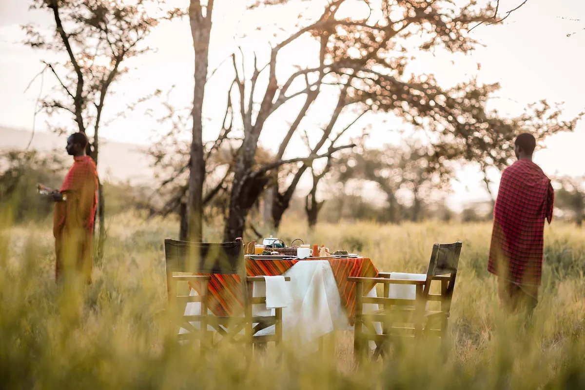 A breakfast table laid in the long grass among acacias with Maasai hosts standing by
