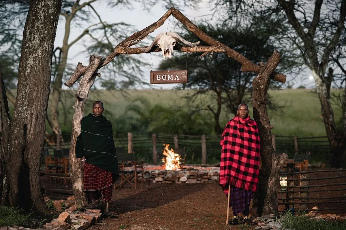 Two Maasai hosts at the entrance to the fire boma at Ole Serai Turner Springs, with the evening fire lit behind them