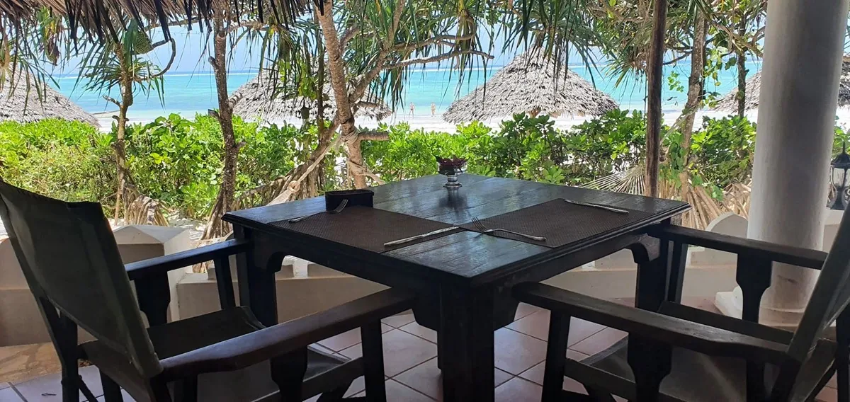 A laid table on the shaded restaurant terrace at Zoi Retreat, looking through palms to the beach and turquoise water